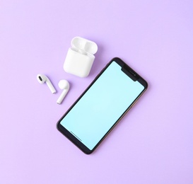 Photo of Wireless earphones, mobile phone and charging case on violet background, flat lay. Space for text