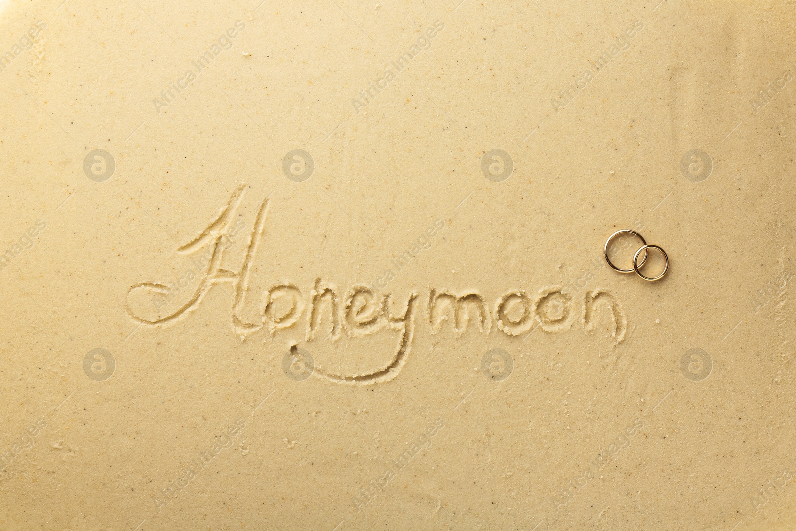 Photo of Word Honeymoon written on sand and two golden rings, top view