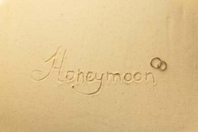 Word Honeymoon written on sand and two golden rings, top view