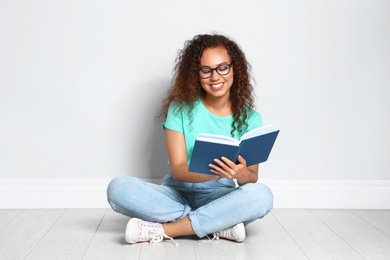 Beautiful young African-American woman reading book on wooden floor near light wall