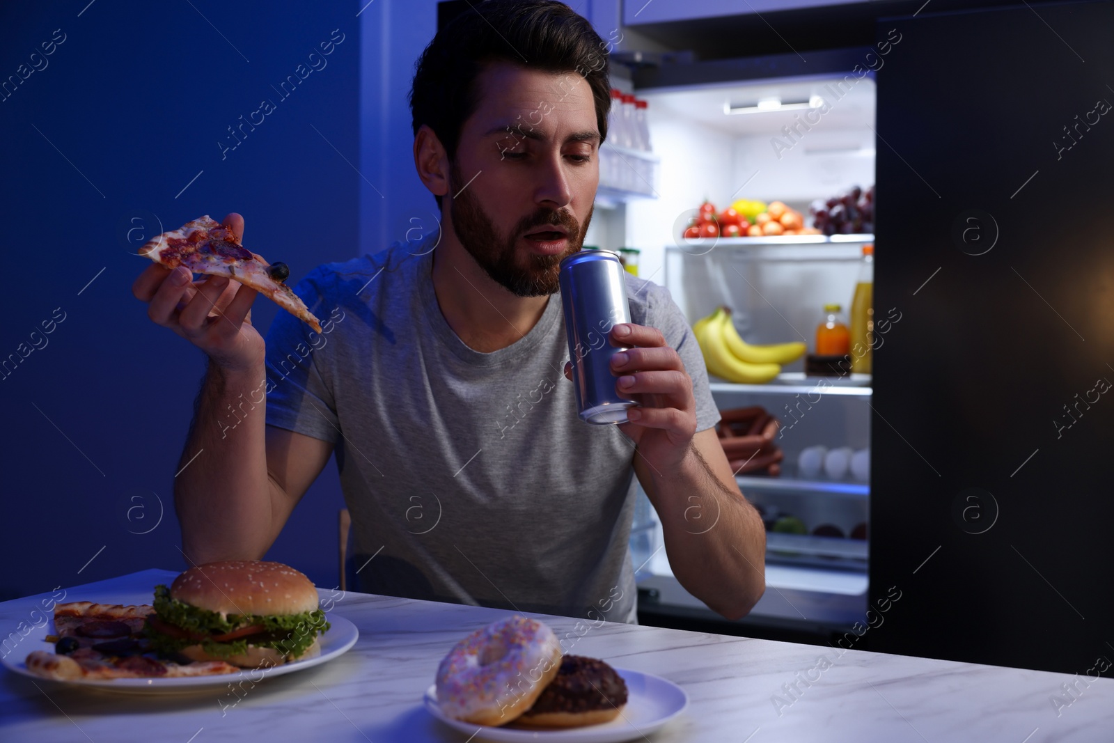 Photo of Man drinking soda and eating junk food in kitchen at night. Bad habit