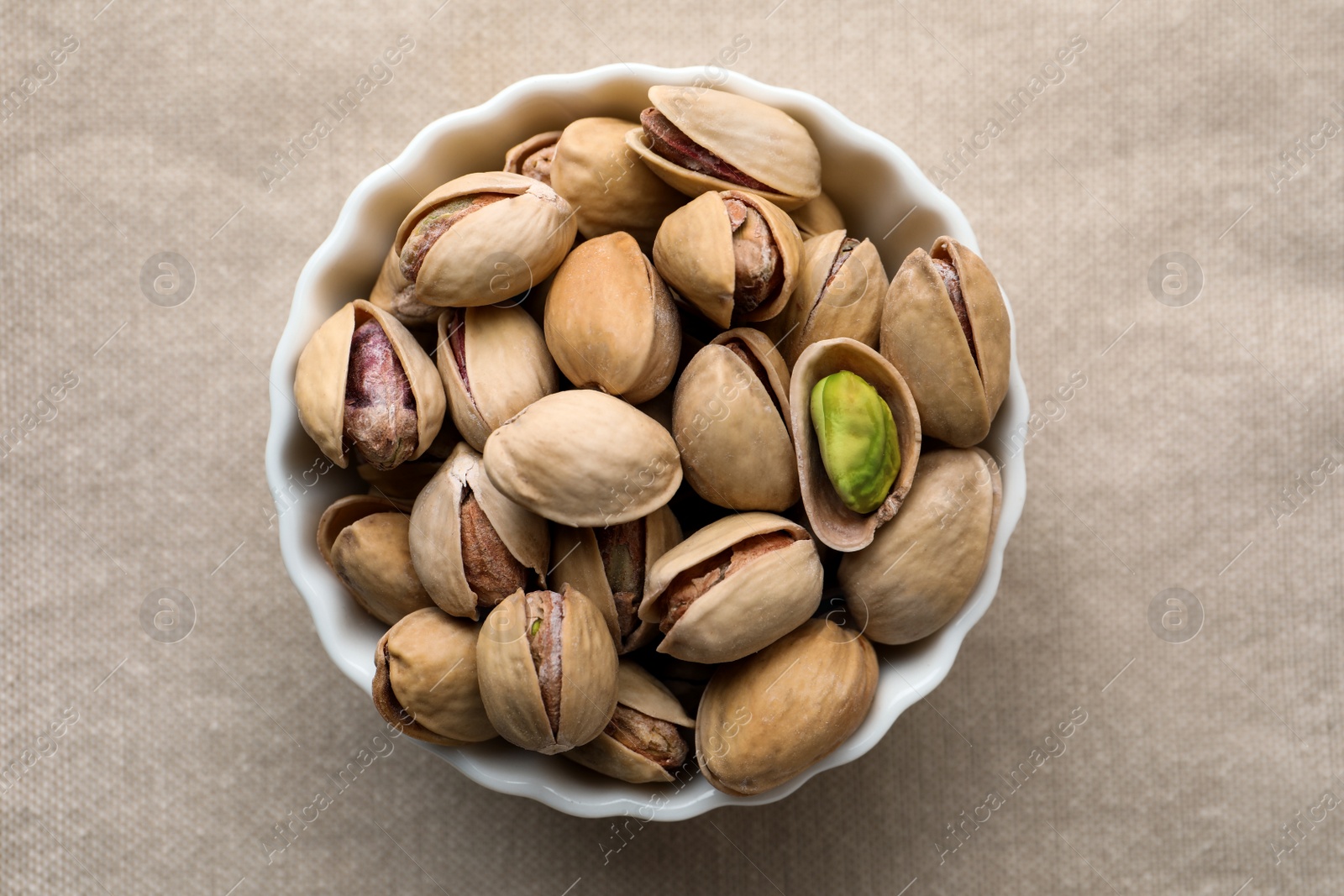 Photo of Bowl with pistachio nuts on beige tablecloth, top view