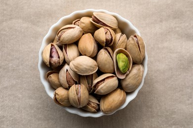 Photo of Bowl with pistachio nuts on beige tablecloth, top view