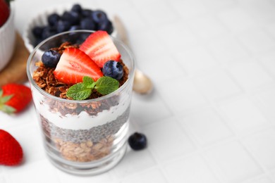 Photo of Tasty granola with berries, yogurt and chia seeds in glass on white tiled table, closeup. Space for text