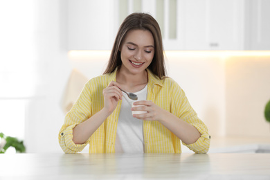 Photo of Young attractive woman eating tasty yogurt at table in kitchen