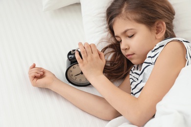 Photo of Cute little girl with alarm clock sleeping in bed, above view