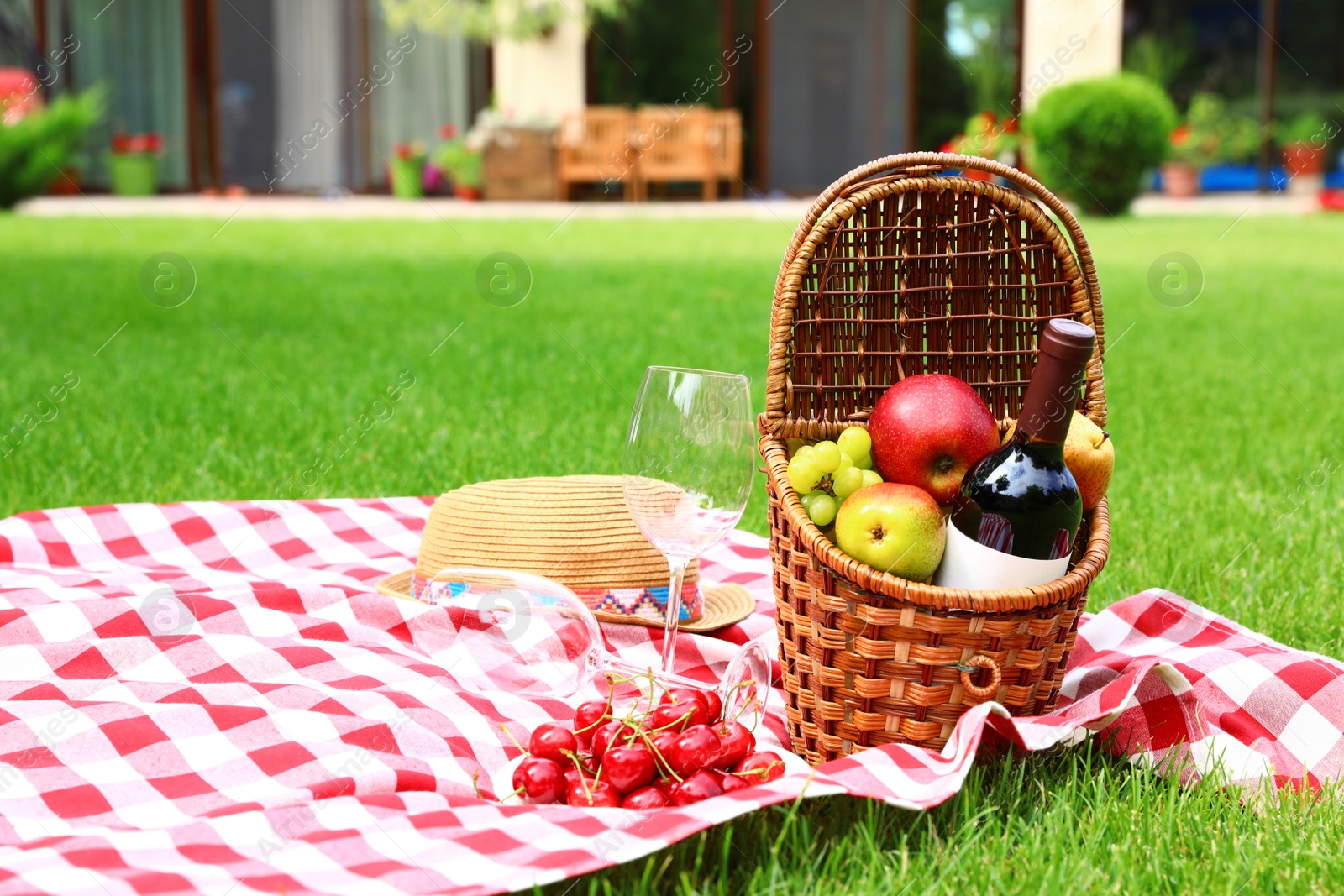 Photo of Picnic basket with fruits and bottle of wine on checkered blanket in garden