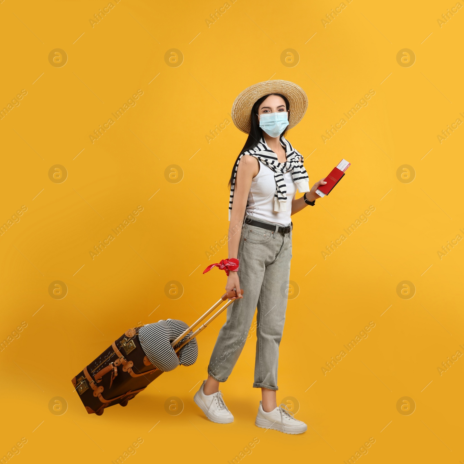 Photo of Female tourist in medical mask with suitcase, ticket and passport on yellow background. Travelling during coronavirus pandemic