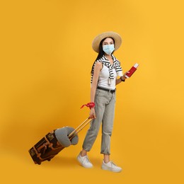 Female tourist in medical mask with suitcase, ticket and passport on yellow background. Travelling during coronavirus pandemic