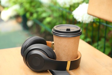 Photo of Paper cup of hot coffee and headphones on wooden bench outdoors. Takeaway drink