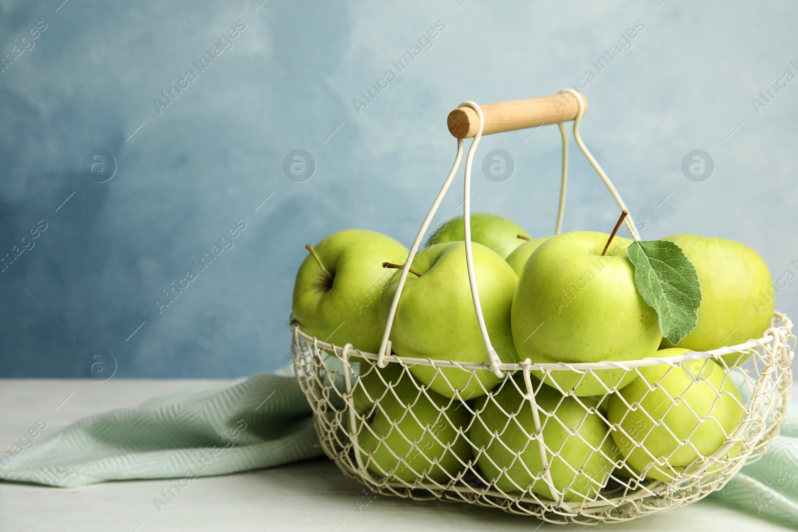 Photo of Metal basket of fresh ripe green apples on white wooden table against blue background, space for text