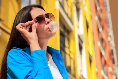 Photo of Beautiful young woman with sunglasses blowing chewing gum on city street, low angle view