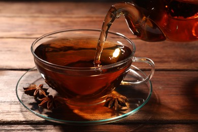 Photo of Pouring aromatic anise tea from teapot into glass cup on wooden table, closeup