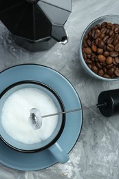 Mini mixer (milk frother), whipped milk in cup, coffee beans and moka pot on grey textured table, flat lay