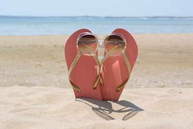 Stylish pink flip flops with sunglasses in sand near sea on sunny day