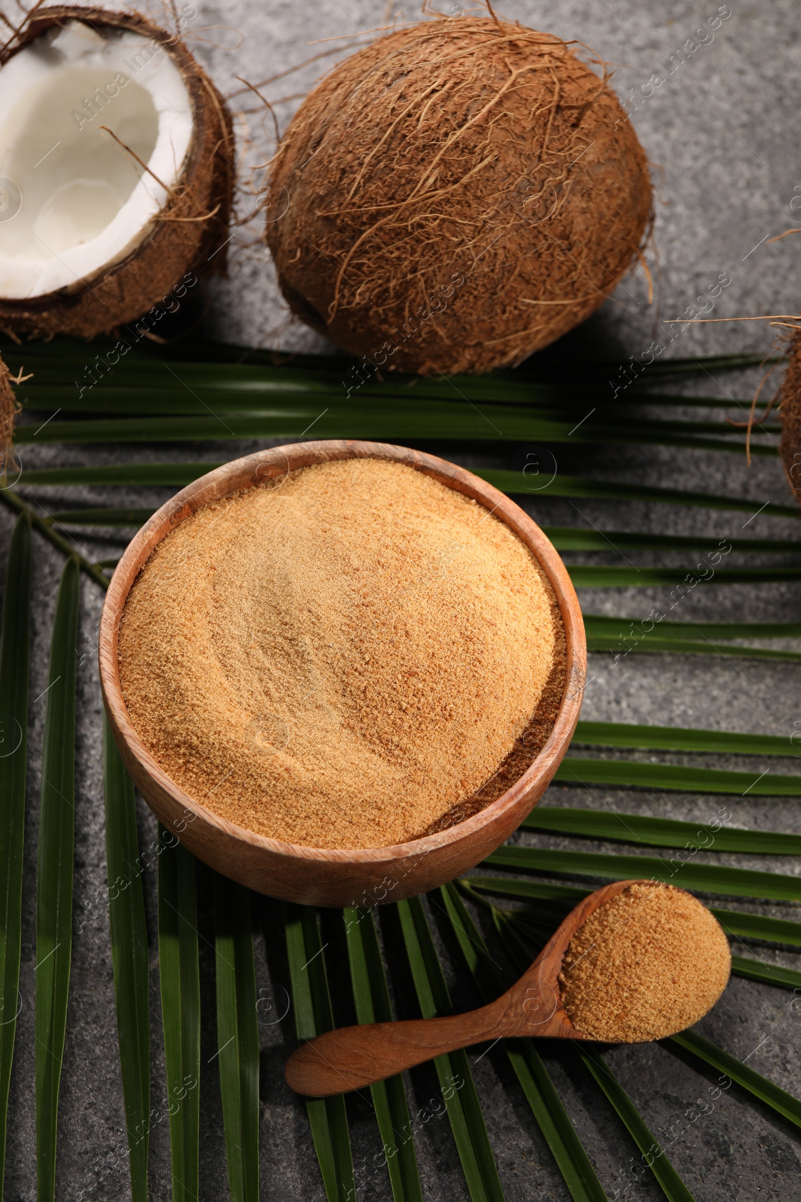 Photo of Coconut sugar in bowl, spoon, palm leaves and fruits on dark textured table, above view