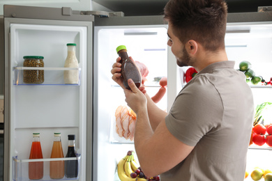 Photo of Young man with bottle of sauce near open refrigerator indoors