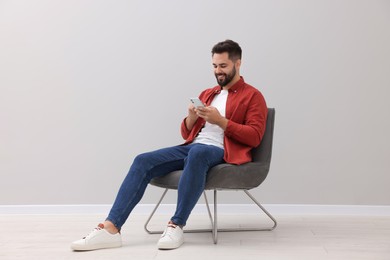 Handsome man using smartphone while sitting in armchair near light grey wall indoors