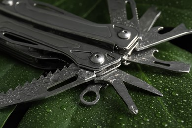 Photo of Compact portable metallic multitool with water drops on green leaf, closeup
