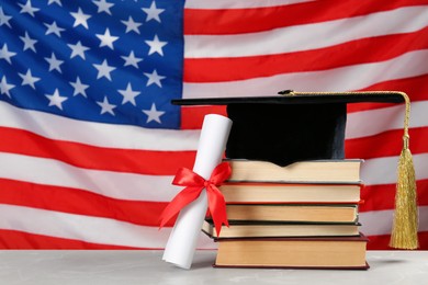 Photo of Graduation hat, books and diploma on light grey table against flag of United States. Space for text