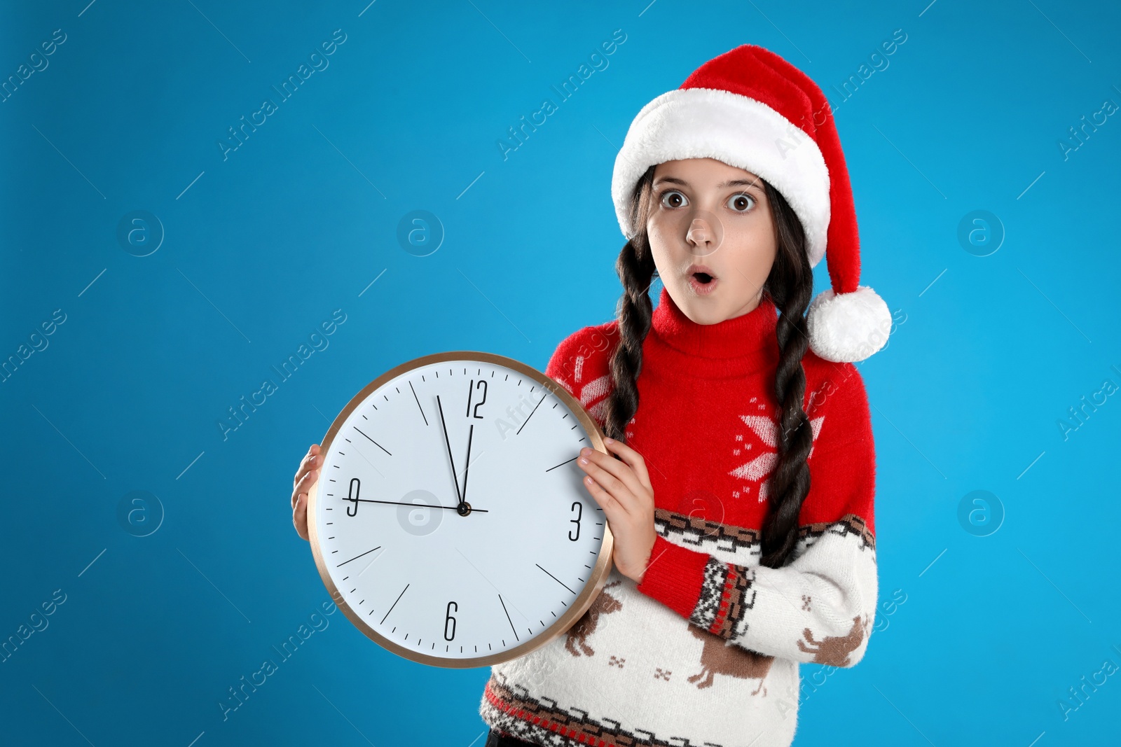 Photo of Girl in Santa hat with clock on light blue background. Christmas countdown