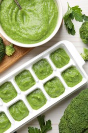Photo of Broccoli puree in ice cube tray and ingredients on white wooden table, flat lay. Ready for freezing