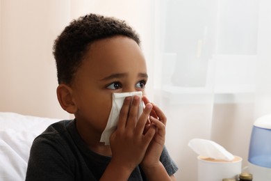 Photo of African-American boy with tissue blowing nose in bed indoors, space for text. Cold symptoms