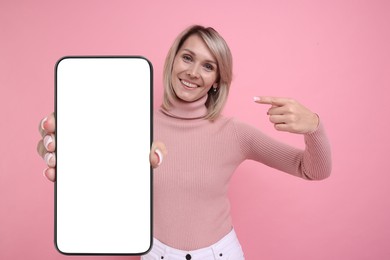 Happy woman pointing at mobile phone with blank screen on pink background. Mockup for design