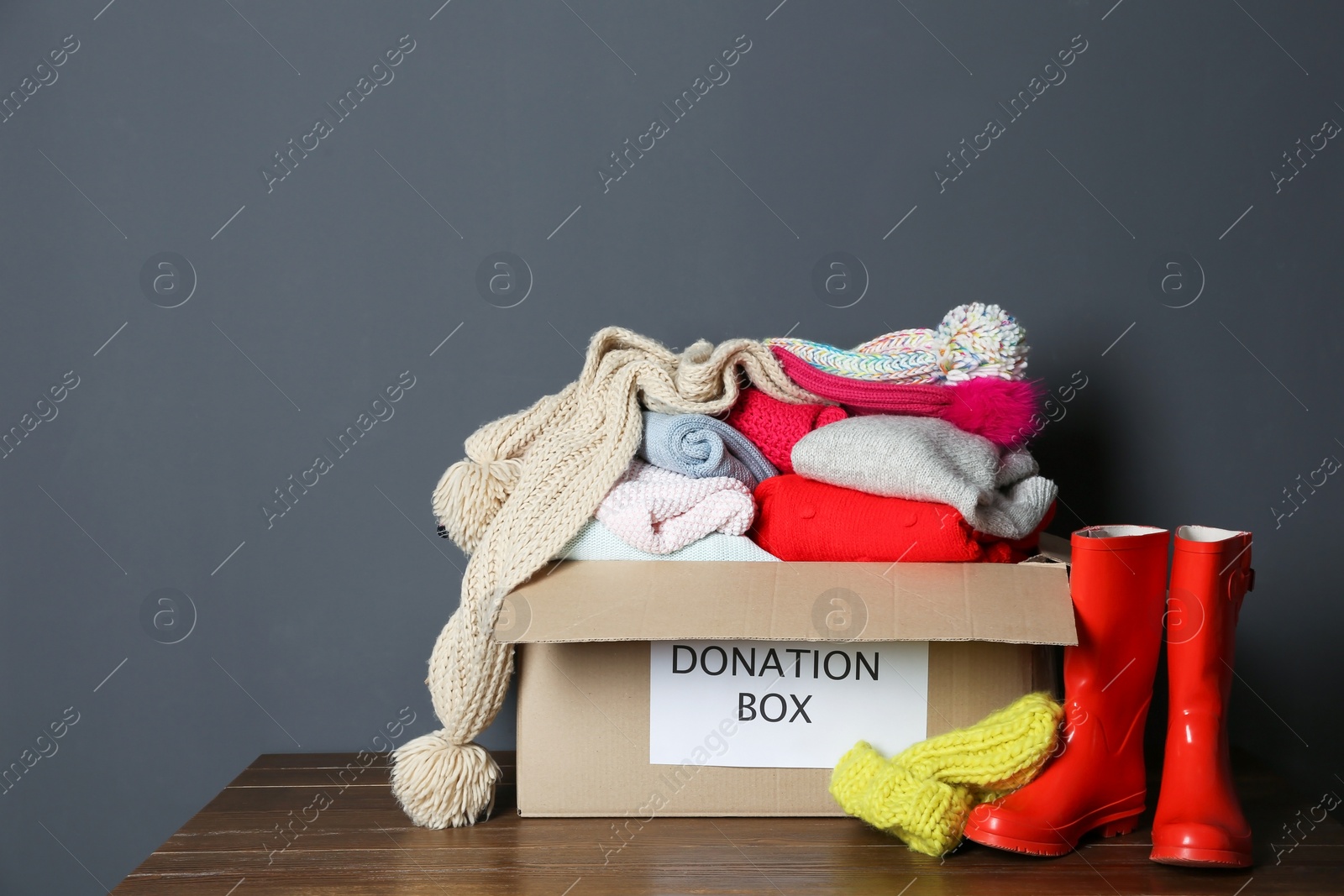Photo of Donation box with knitted clothes and rubber boots on table near grey wall. Space for text