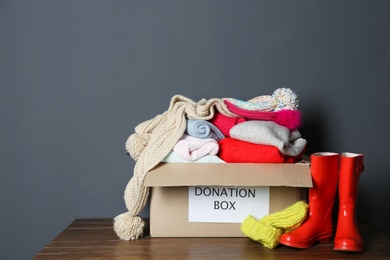 Photo of Donation box with knitted clothes and rubber boots on table near grey wall. Space for text