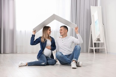 Photo of Young family housing concept. Pregnant woman with her husband sitting under cardboard roof on floor at home