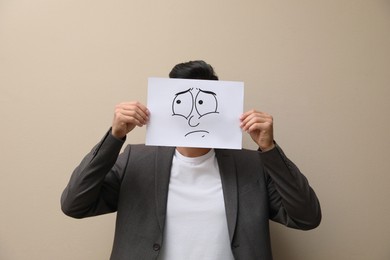 Photo of Man hiding emotions using card with drawn frowning face on beige background