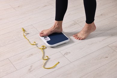 Woman stepping on floor scale and measuring tape at home, closeup. Weight control