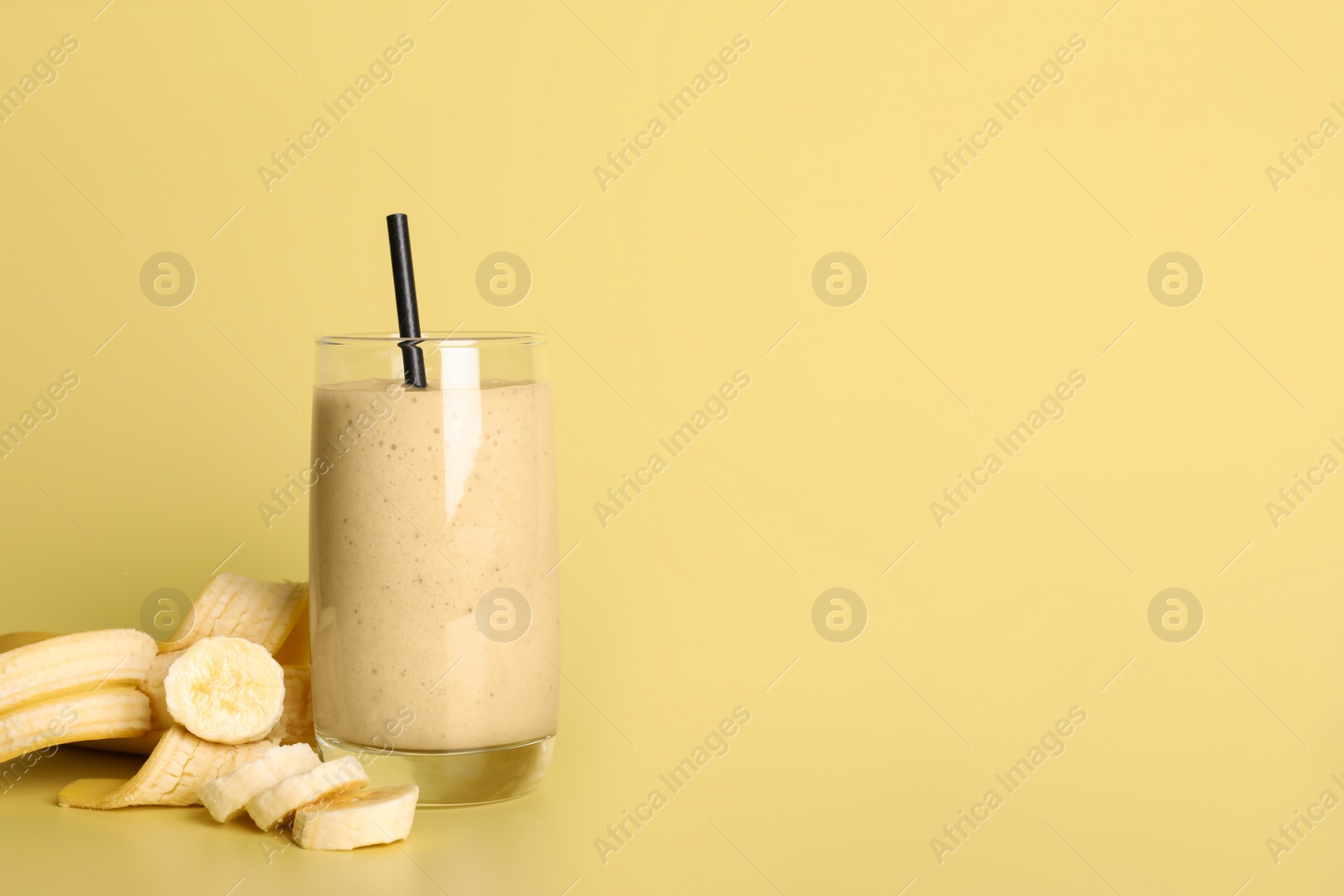 Photo of Glass of tasty smoothie with straw and cut banana on pale yellow background. Space for text