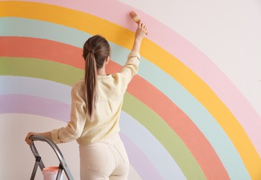 Young woman painting rainbow on white wall indoors, back view. Space for text