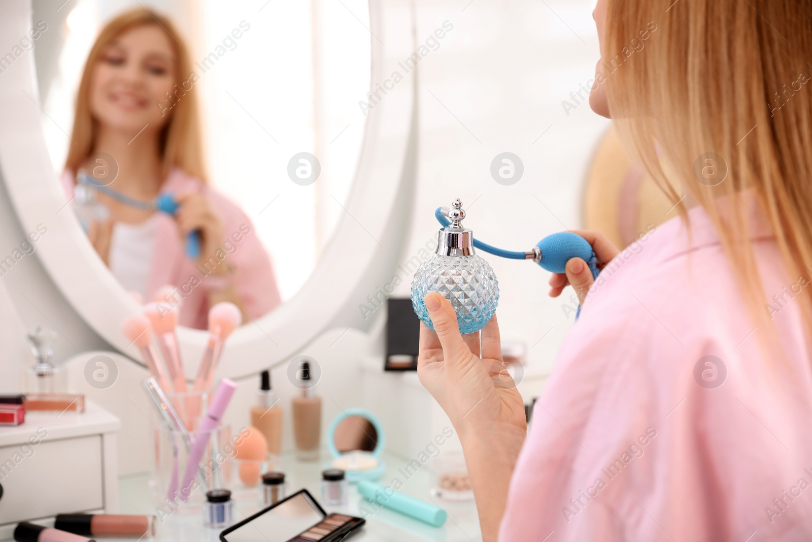 Photo of Young woman with beautiful makeup applying perfume near mirror indoors