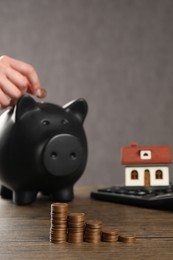 Woman putting coin into piggy bank, closeup. House model, calculator and money on wooden table, selective focus