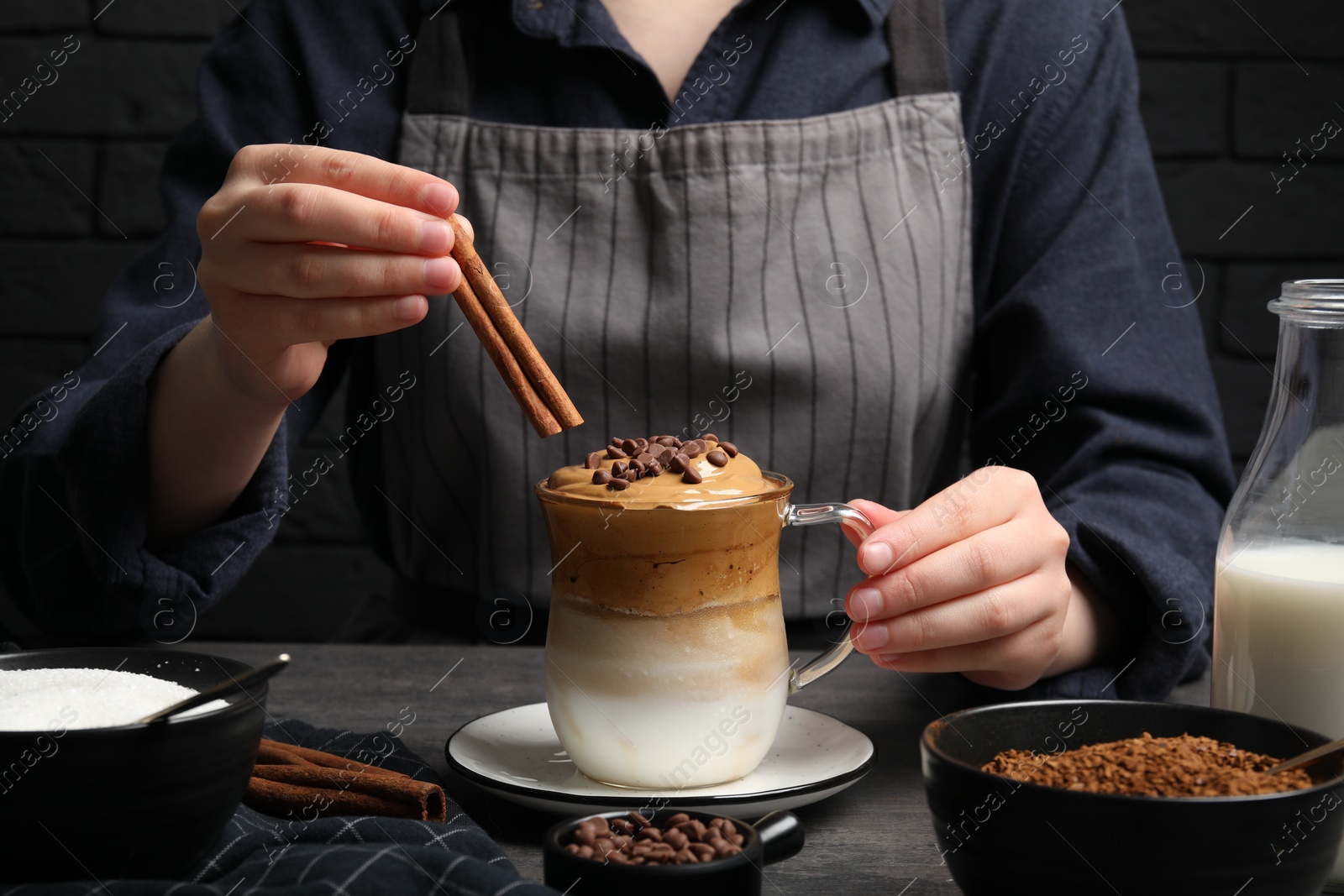 Photo of Woman decorating dalgona coffee with cinnamon sticks at grey table, closeup