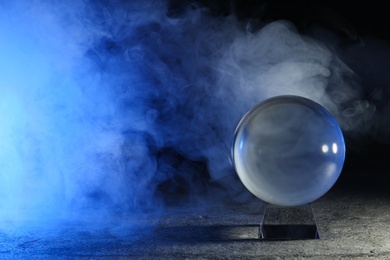 Photo of Magic crystal ball  on grey table and smoke against dark background, space for text. Making predictions