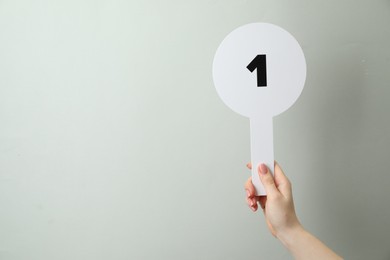 Woman holding auction paddle with number 1 on light grey background, closeup. Space for text