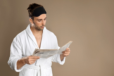 Photo of Young man in bathrobe and eye sleeping mask  reading newspaper on brown background. Space for text