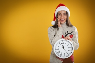 Woman in Santa hat with clock on yellow background, space for text. Christmas countdown