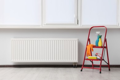 Photo of Ladder with cleaning supplies near modern radiator in room