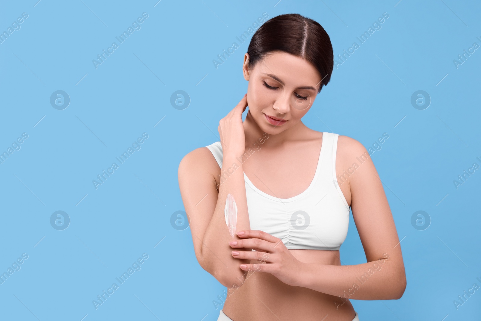 Photo of Beautiful woman with smear of body cream on her arm against light blue background, space for text