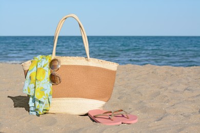 Photo of Straw bag with beach wrap, sunglasses and flip flops on sandy seashore, space for text. Summer accessories