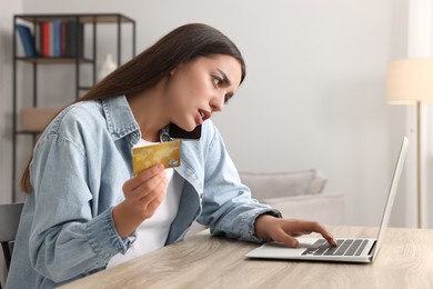 Confused woman with credit card talking on phone and using laptop at table indoors. Be careful - fraud