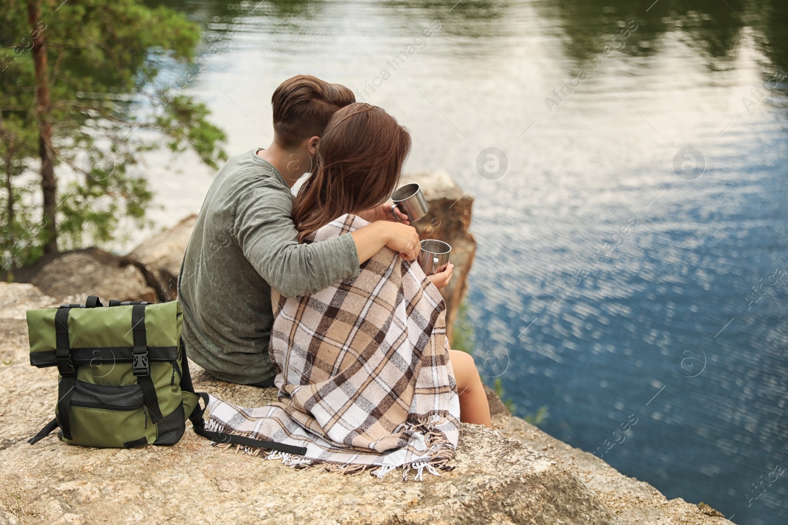 Photo of Cute couple with mugs and plaid near lake. Camping season