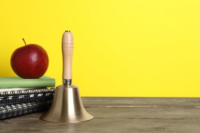 Photo of Golden school bell, apple and notebooks on wooden table against yellow background, space for text
