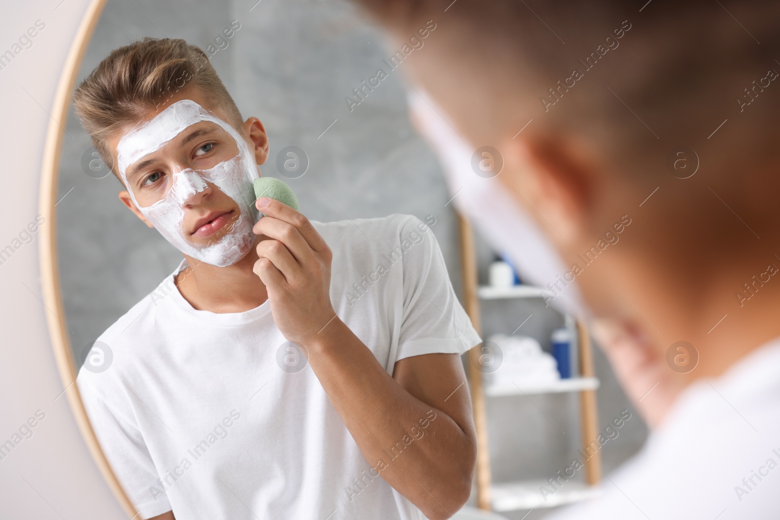 Photo of Young man washing off face mask with sponge near mirror in bathroom