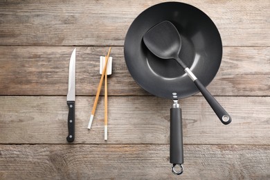 Photo of Black metal wok, chopsticks, knife and spatula on wooden table, flat lay